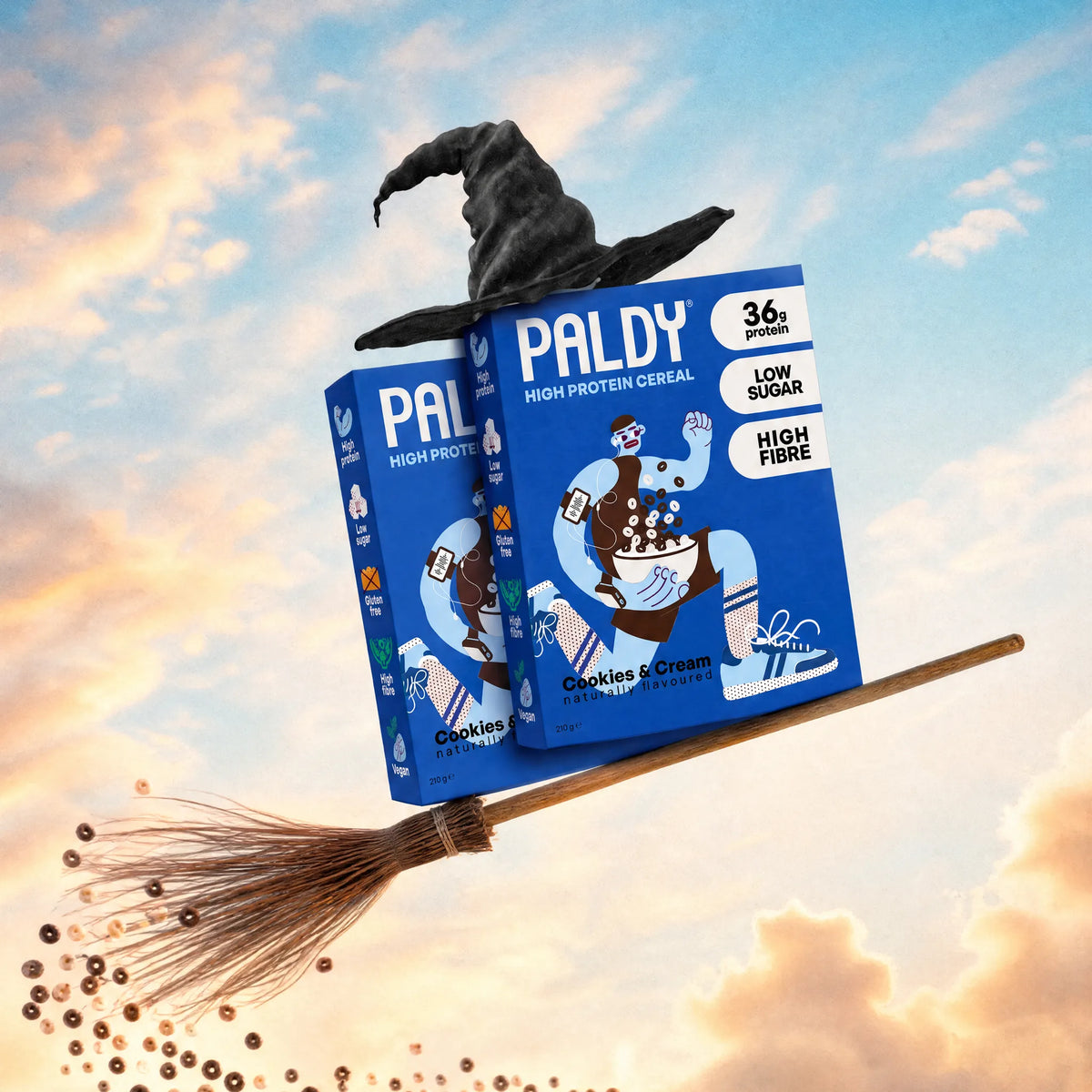 Two Paldy Cookies & Cream high protein cereal boxes wearing a witch hat, flying on a broom through a sunset sky with a trail of cereal